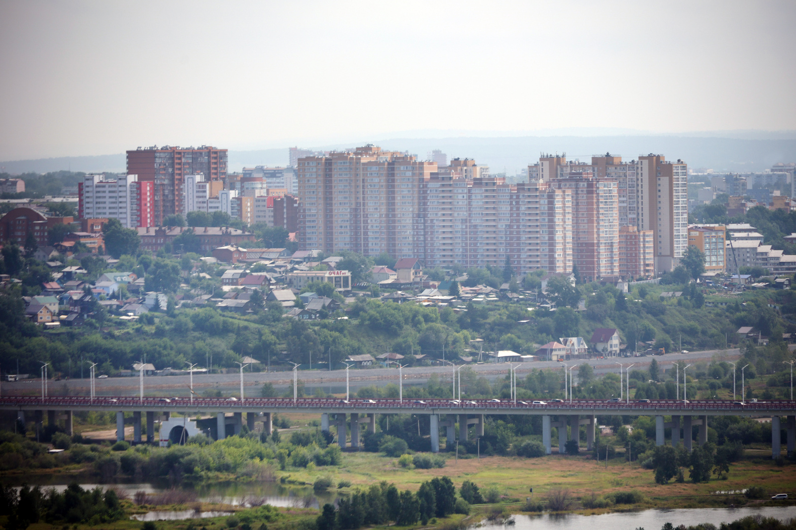 Мост через реку и городской пейзаж с высотками под серым небом.