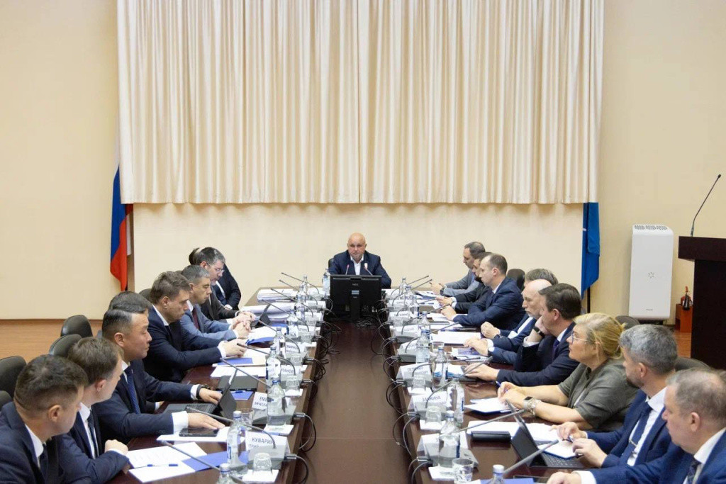 Люди на заседании с флагами России на стене.